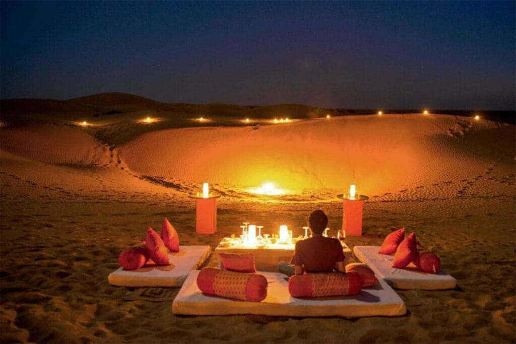 a man sitting on a bed in the desert with a lit fire
